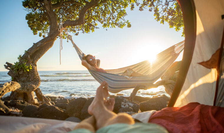 Point of view of man's feet from inside a tent camping on the beach in Hawaii looking at girlfriend in hammock outdoors