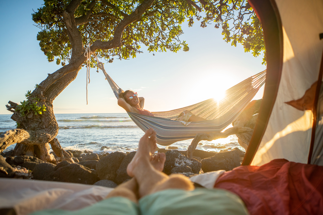 Point of view of man's feet from inside a tent camping on the beach in Hawaii looking at girlfriend in hammock outdoors